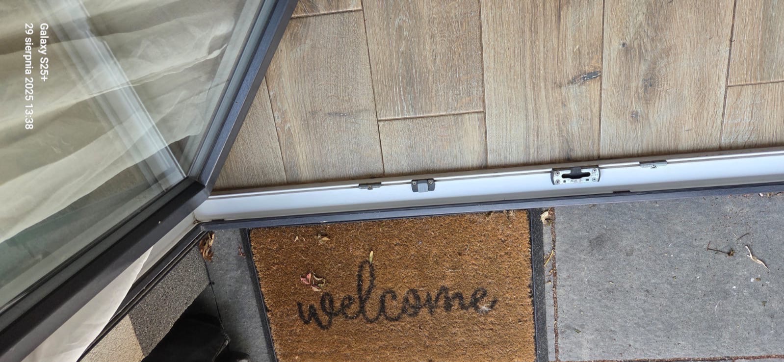 Otwarty balkon z drewnianą podłogą i wycieraczką 'welcome' na szarym betonie. Widoczna rama okienna i zasłona w tle. Ujęcie z góry.