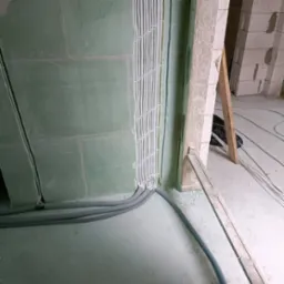 Nowa instalacja elektryczna: równolegle ułożone przewody w białych rurkach mocowane do zielonej ściany w surowym stanie, z szarymi rurami karbowanymi na podłodze i widoczną konstrukcją budynku w tle.