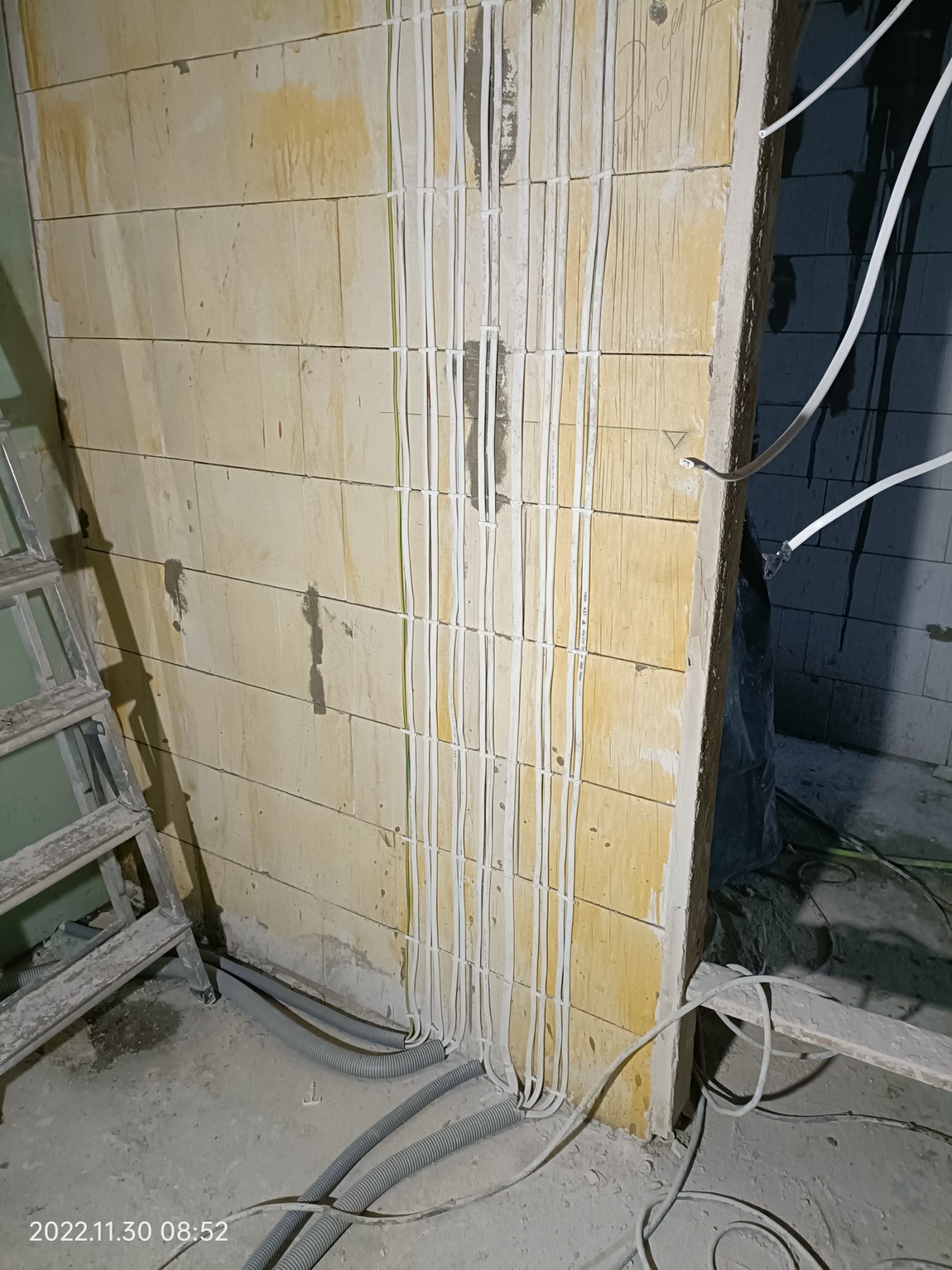 Montaż przewodów elektrycznych w nowym budynku, widoczne kable przymocowane do ściany, drabina i rury karbowane na podłodze w trakcie prac wykończeniowych.