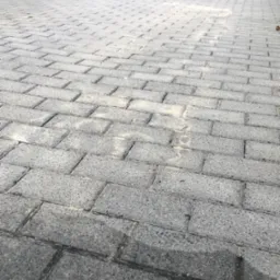 Szara kostka brukowa na podjeździe, częściowo umyta, z widocznymi śladami piasku w spoinach, prowadząca do metalowej bramy.