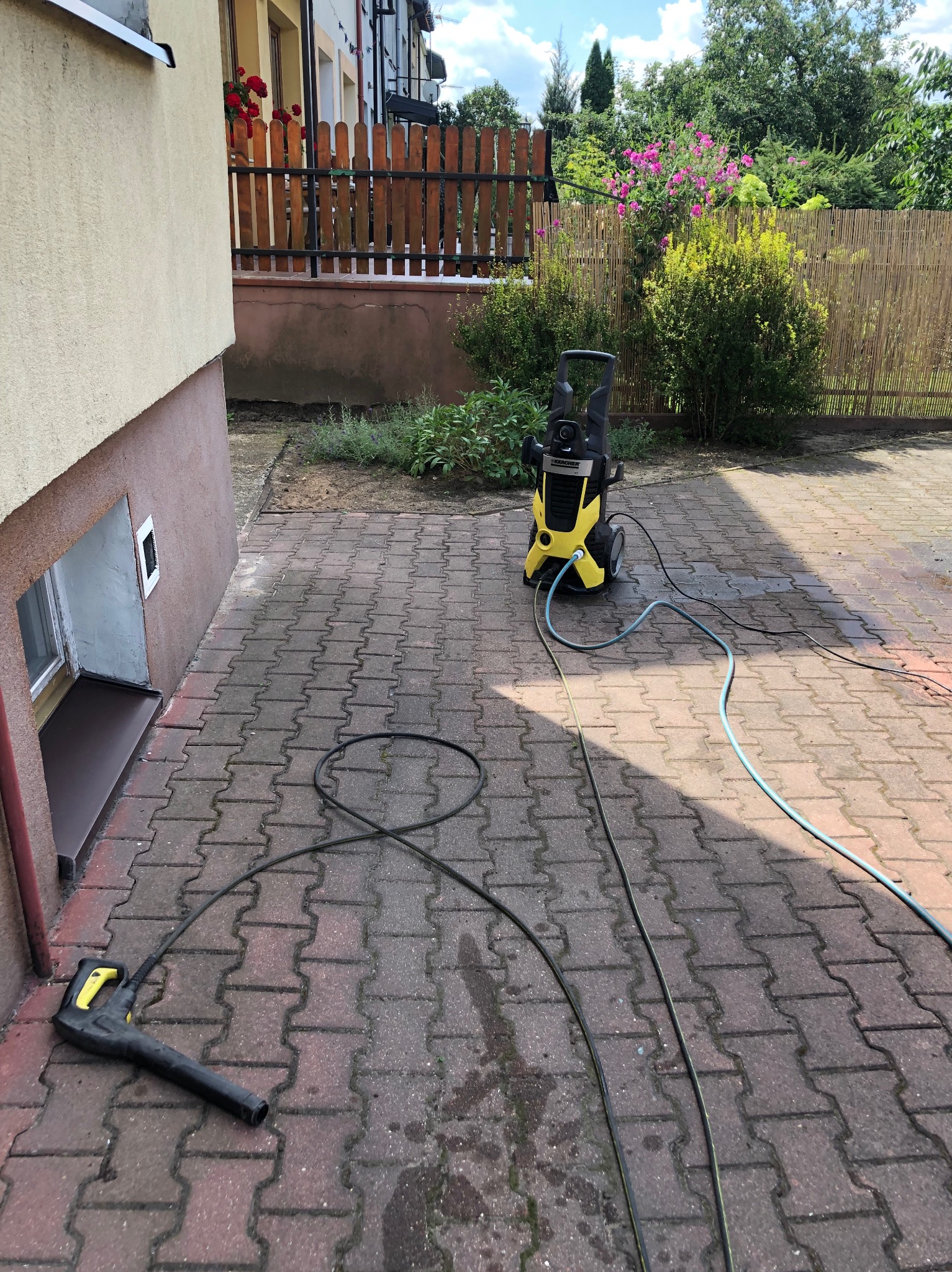 Żółto-czarna myjka ciśnieniowa Kärcher na kostce brukowej, z widocznym pistoletem natryskowym i wężami, częściowo mokra powierzchnia kostki po czyszczeniu.