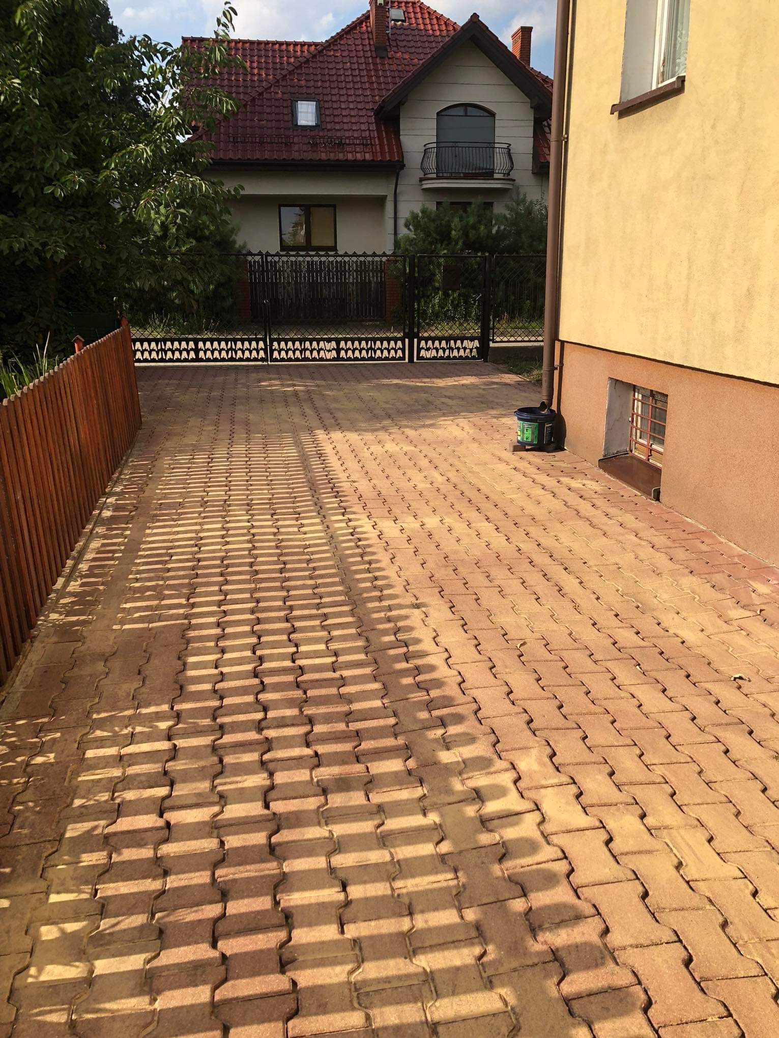 Długa, wyłożona brązowo-żółtą kostką brukową droga dojazdowa, oświetlona promieniami słońca, z widocznym fragmentem domu i drewnianym płotem po lewej stronie oraz ścianą budynku po prawej stronie.