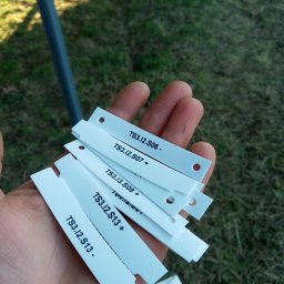 ElektroComplex - Dłoń trzymająca białe, plastikowe oznaczniki kablowe z nadrukowanymi numerami TS3.I2.S06, TS3.I2.S07, TS3.I2.S08 i TS3.I2.S13, z otworami i wycięciami na kable, na tle trawy i metalowej konstrukcji.