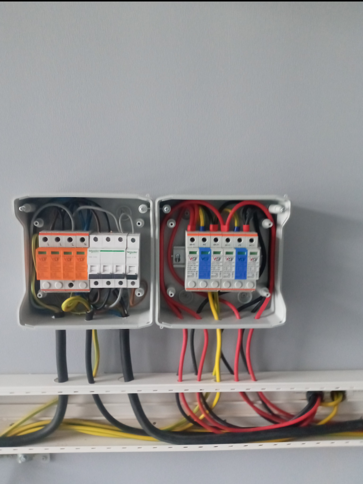Dwa otwarte panele elektryczne z różnymi zabezpieczeniami, okablowaniem w kolorach czerwonym, czarnym i żółtym, zamontowane na białej listwie przypodłogowej przy szarej ścianie.