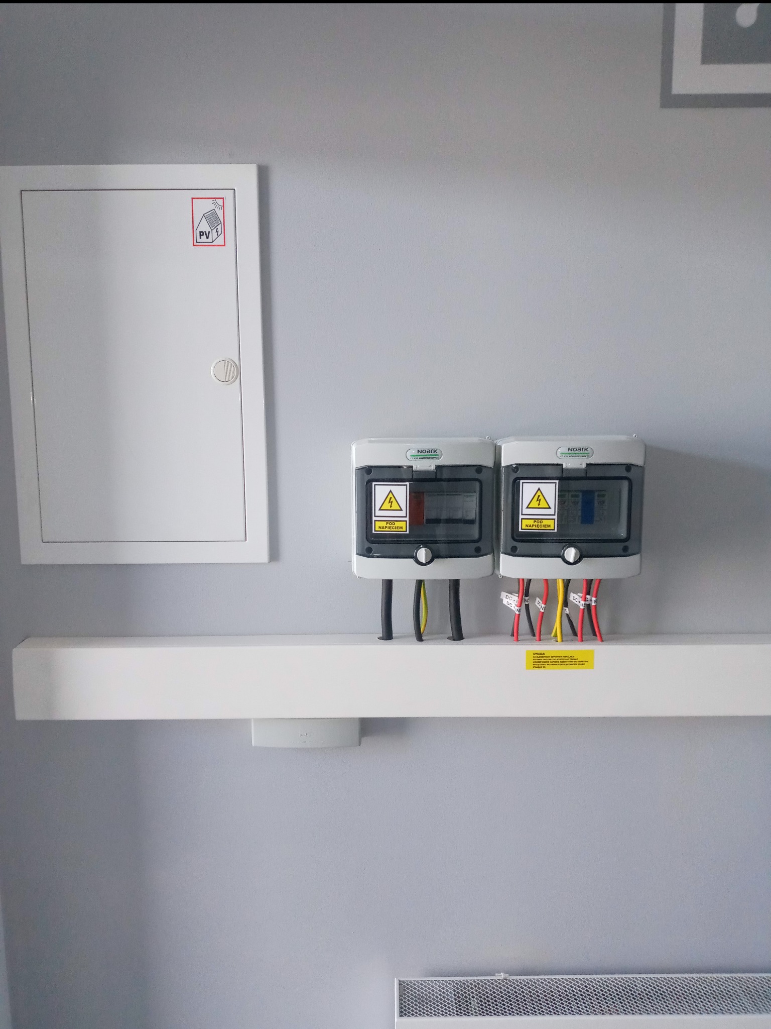 Dwa szare zabezpieczenia nadprądowe DC firmy Noark na białej półce, z podłączonymi przewodami o różnych kolorach, zamontowane na szarej ścianie obok białej skrzynki z symbolem fotowoltaiki.