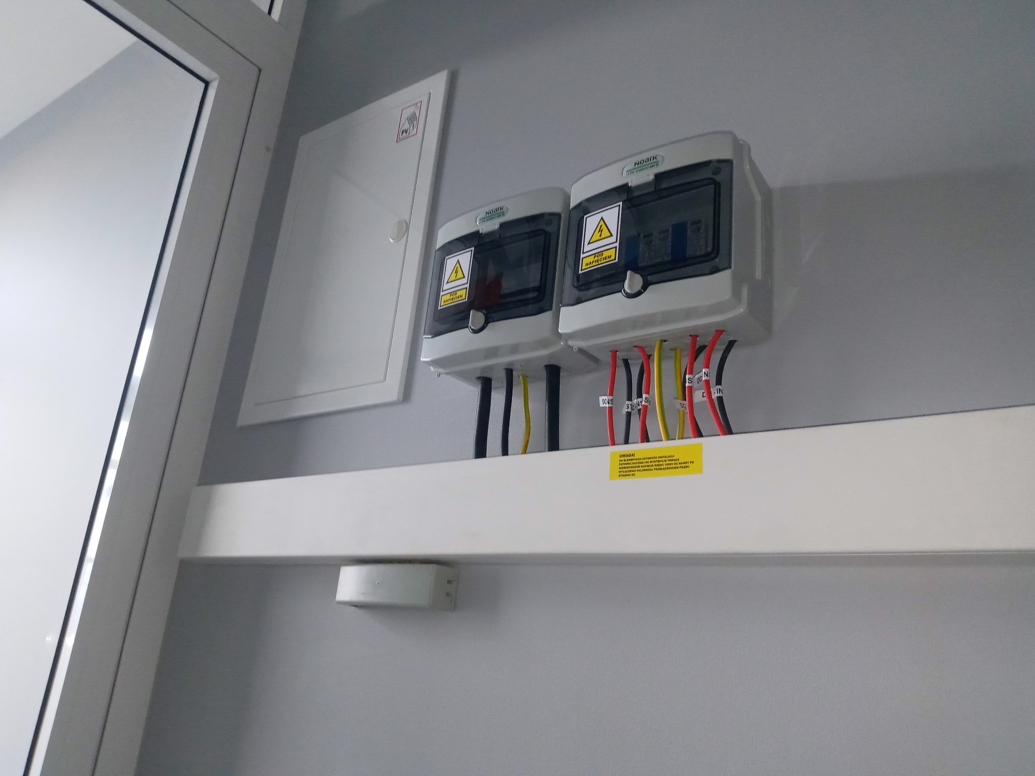Dwie skrzynki elektryczne Noark z ostrzeżeniem o napięciu, zamontowane na ścianie, z widocznymi przewodami o różnych kolorach i żółtą naklejką informacyjną poniżej. Obok widoczna biała skrzynka PV.