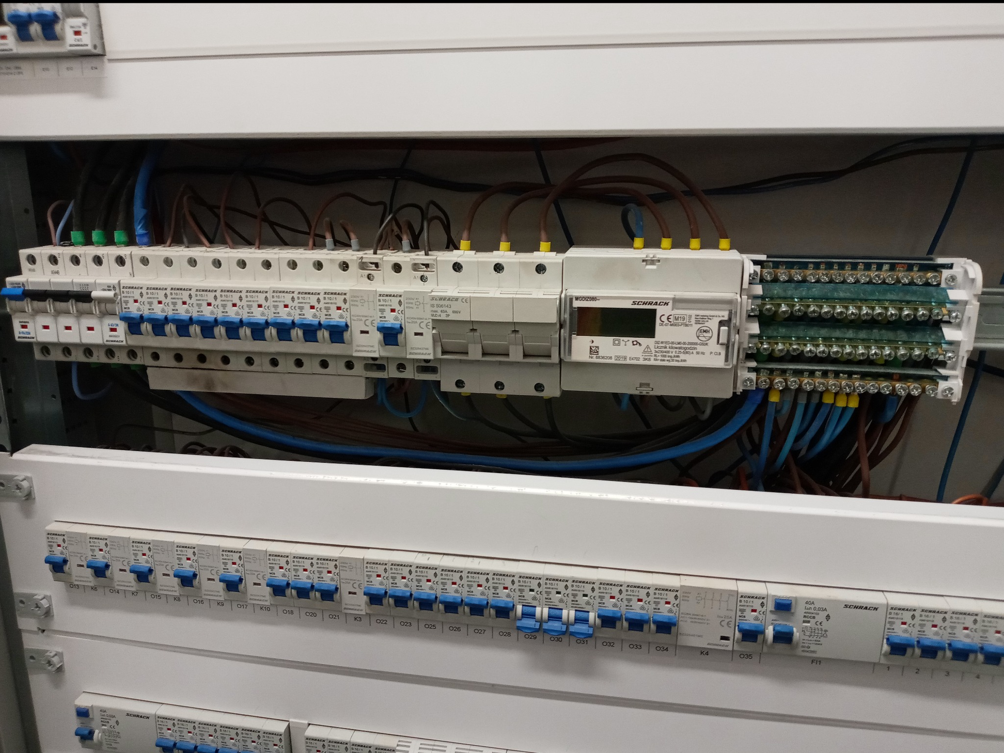 Rozdzielnica elektryczna Schrack z zamontowanymi bezpiecznikami i licznikiem energii elektrycznej, widoczne przewody i oznaczenia.