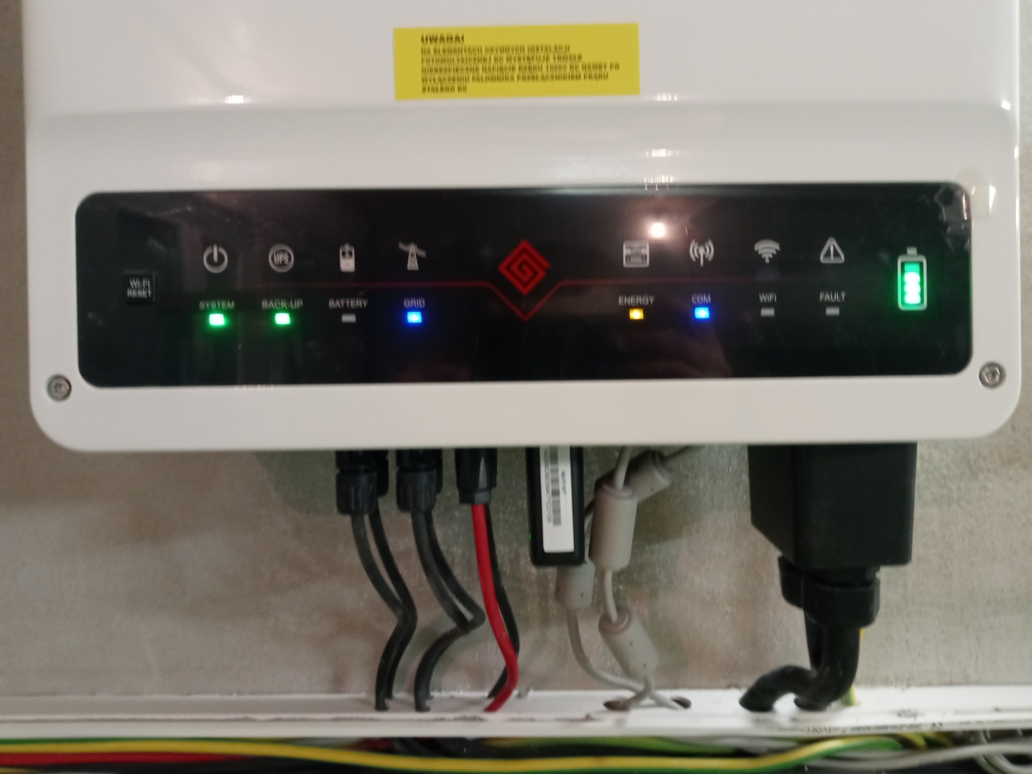 Biała obudowa inwertera fotowoltaicznego z widocznymi diodami LED wskazującymi status pracy, z podłączonymi kablami zasilającymi i komunikacyjnymi.