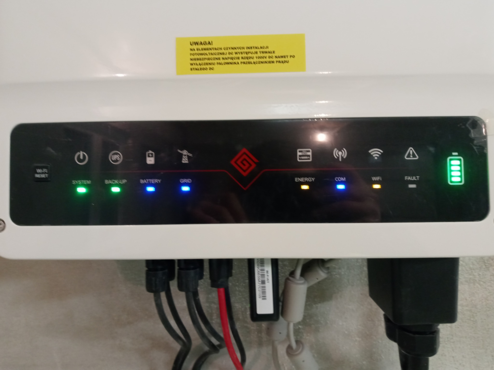 Panel sterowania inwertera fotowoltaicznego z diodami LED wskazującymi status pracy systemu, baterii, sieci i komunikacji Wi-Fi; widoczne kable zasilające i naklejka ostrzegawcza o wysokim napięciu.