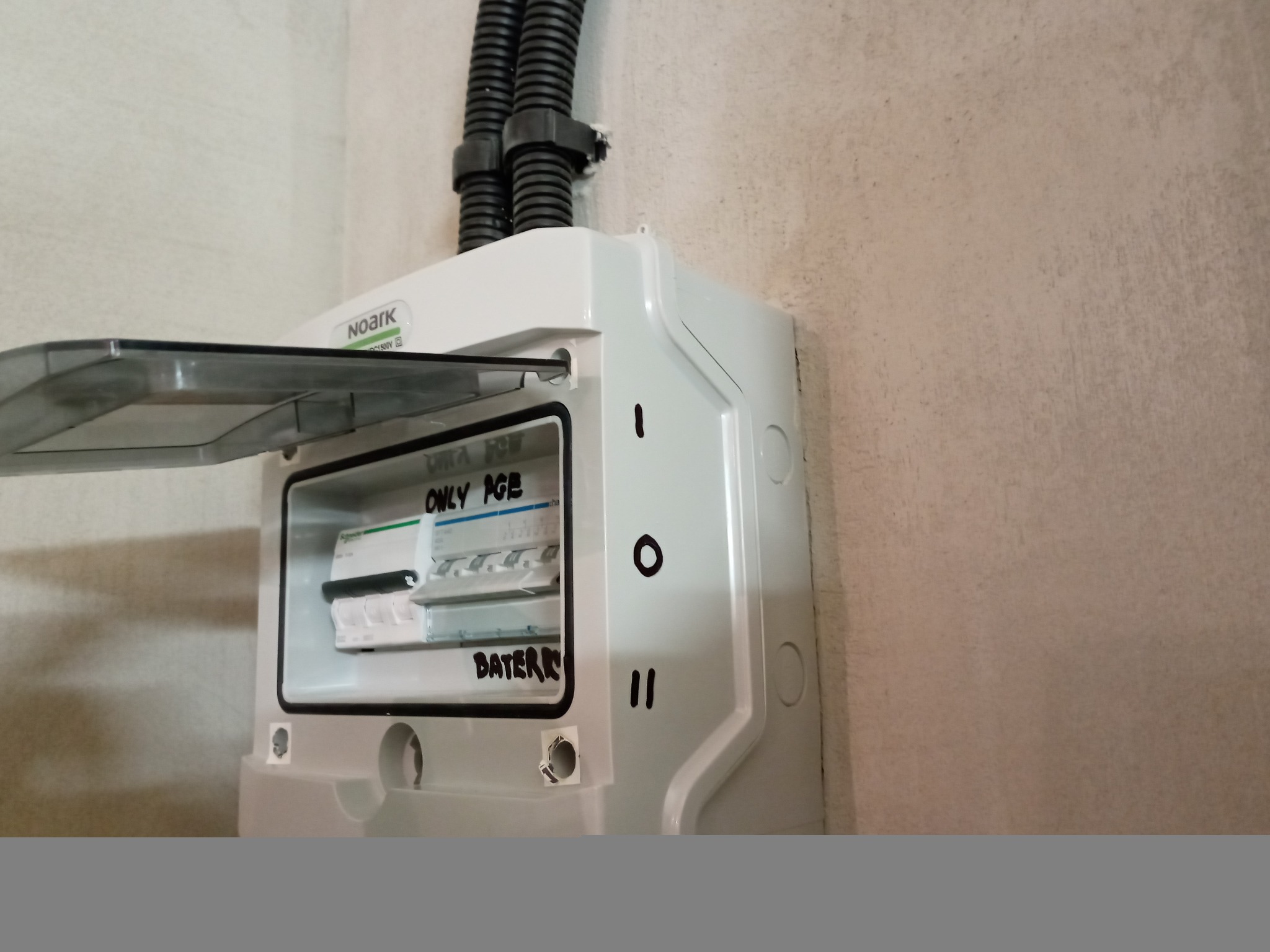 Otwarta skrzynka elektryczna Noark z zabezpieczeniami, widoczne napisy markerem 'ONLY PGE' i 'BATERIE', kable w peszlu wchodzące od góry.