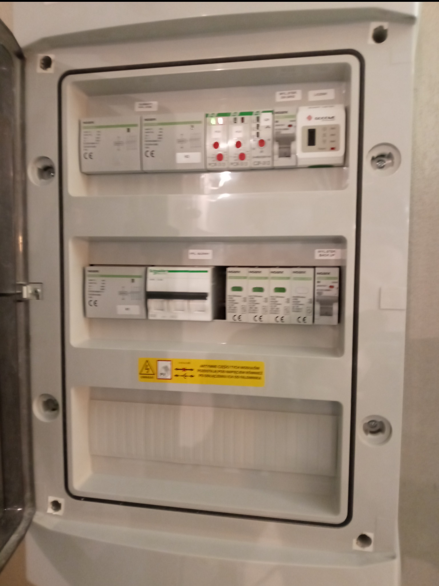 Otwarta skrzynka elektryczna z zainstalowanymi zabezpieczeniami przepięciowymi, wyłącznikami różnicowoprądowymi i modułem sterującym, widoczna żółta naklejka ostrzegawcza z symbolem wysokiego...