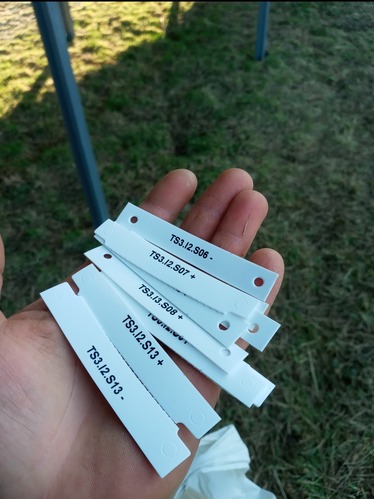 Dłoń trzymająca białe, plastikowe oznaczniki kablowe z nadrukowanymi numerami TS3.I2.S06, TS3.I2.S07, TS3.I2.S08 i TS3.I2.S13, z otworami i wycięciami na kable, na tle trawy i metalowej konstrukcji.