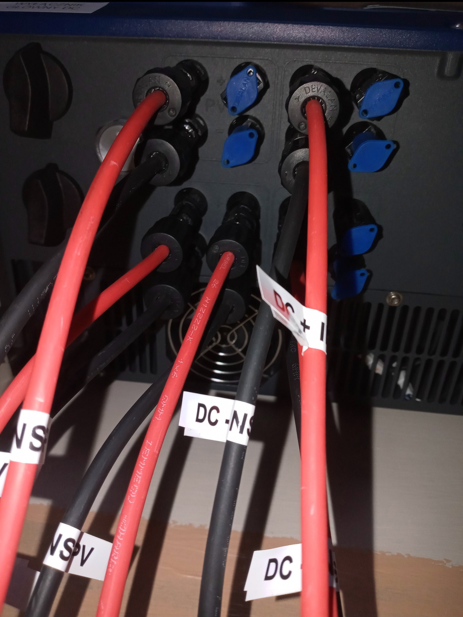 Szczegółowe ujęcie połączeń kabli DC oznaczonych etykietami w falowniku instalacji fotowoltaicznej, widoczne czerwone i czarne przewody z oznaczeniami DC+, DC-NS oraz niebieskie zaślepki.