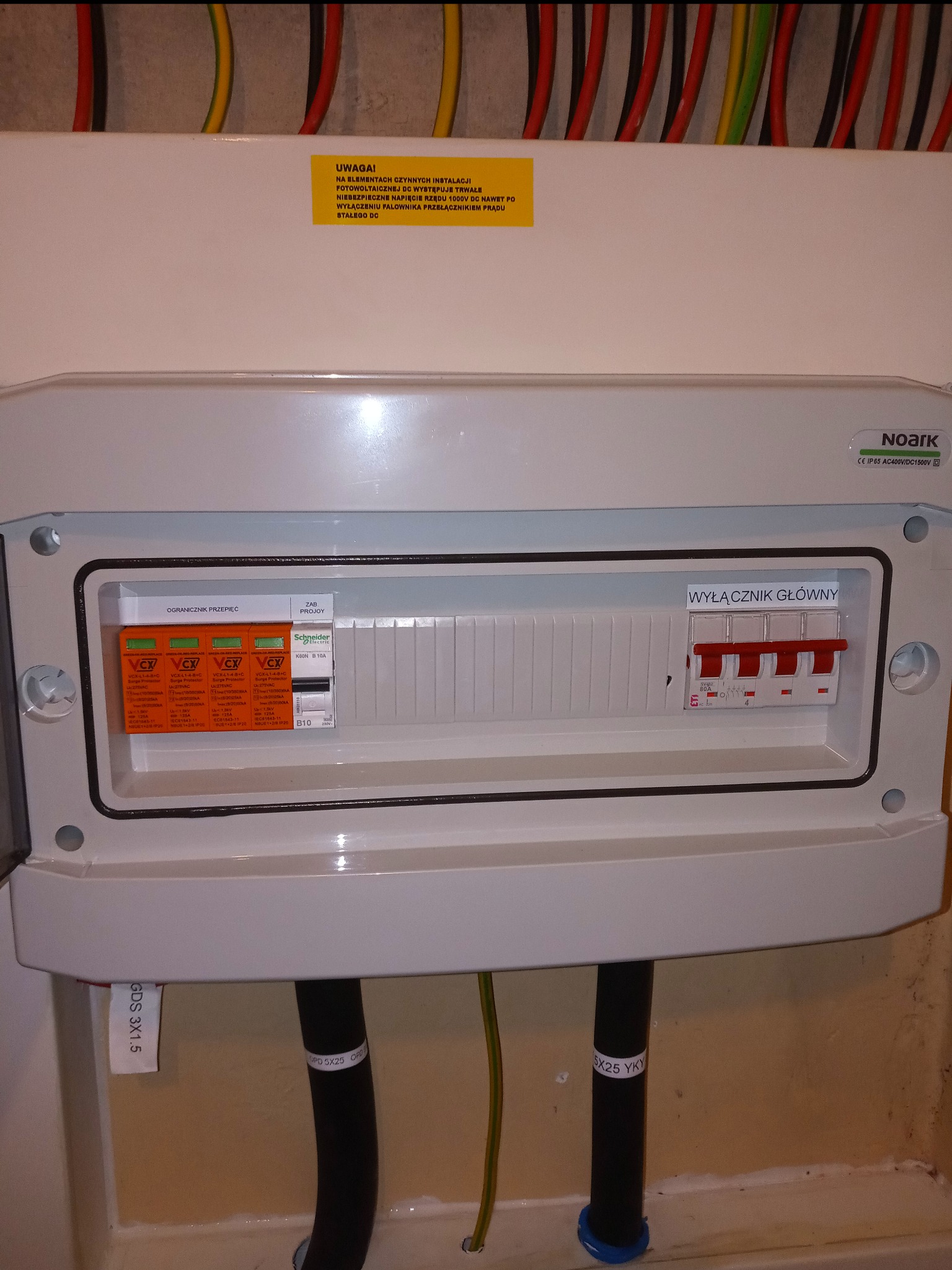 Rozdzielnica elektryczna z wyłącznikiem głównym i ogranicznikami przepięć, z żółtą naklejką ostrzegawczą o wysokim napięciu w instalacji fotowoltaicznej, widoczne kable wchodzące do skrzynki.