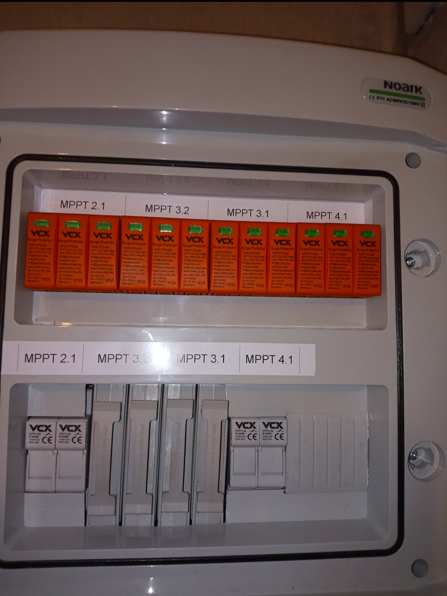 Rozdzielnica fotowoltaiczna Noark z widocznymi bezpiecznikami i ogranicznikami przepięć VOX dla różnych MPPT, etykiety z opisami, widoczne oznaczenia CE i IP65.
