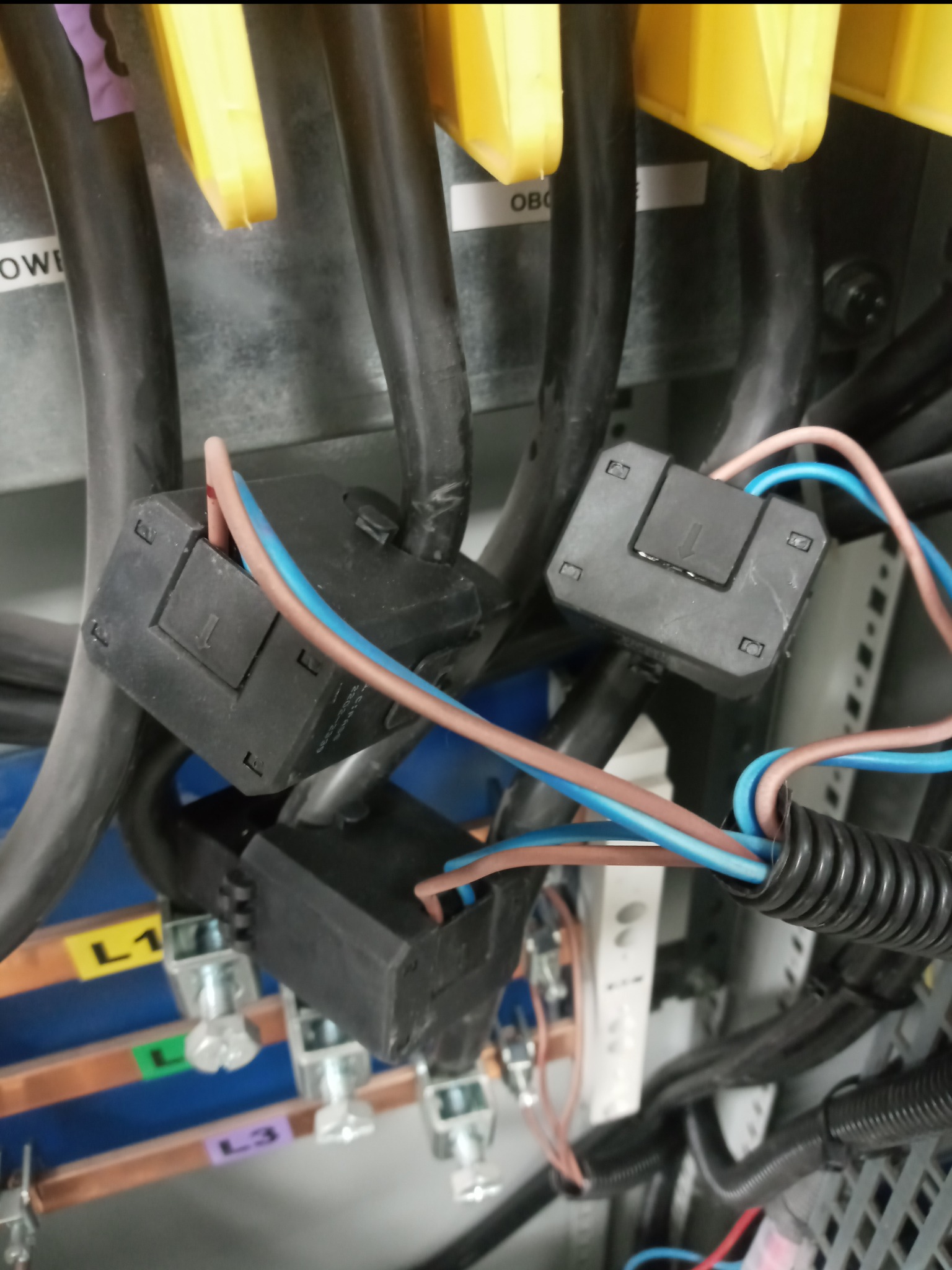 Szczegółowe ujęcie z wnętrza szafy elektrycznej, widoczne czarne kostki łączeniowe z podłączonymi przewodami w kolorach brązowym i niebieskim, oraz grube kable z żółtymi osłonami, z oznaczeniami L1...