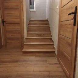 Klatka schodowa z drewnianymi schodami i panelami podłogowymi imitującymi drewno, dwie drewniane drzwi z czarnymi klamkami po bokach, ściana z dekoracyjną szarą fakturą i małe okno na podeście.