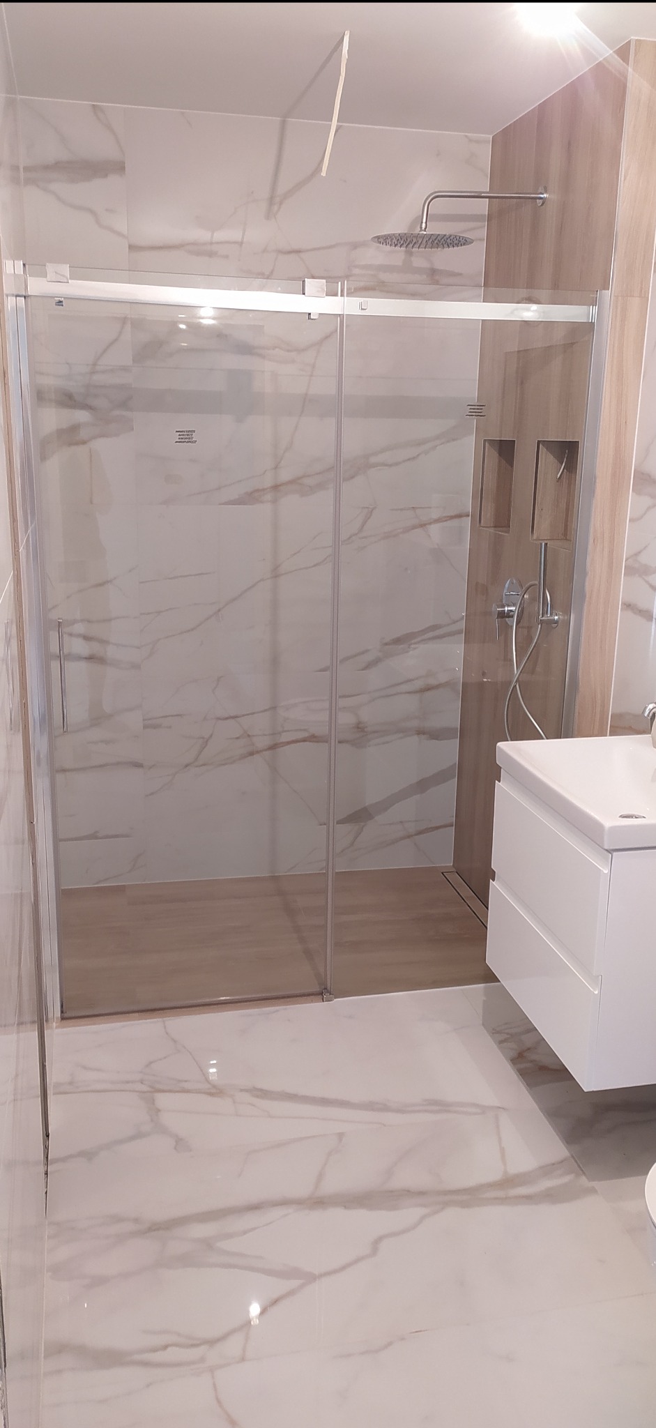 Nowoczesna kabina prysznicowa z przesuwnymi drzwiami, wykończona płytkami imitującymi marmur, z deszczownicą i wnękami na kosmetyki w ścianie.