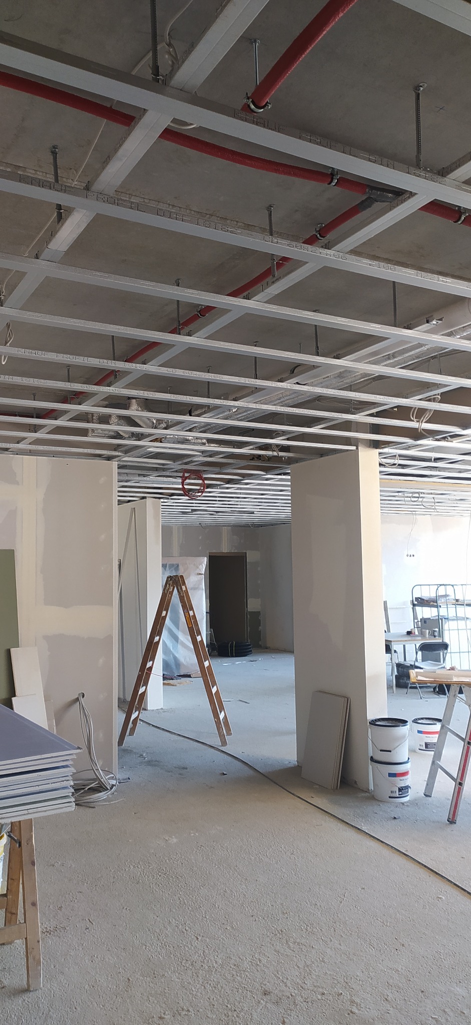 Wnętrze w trakcie prac wykończeniowych: widoczne płyty gipsowo-kartonowe, metalowa konstrukcja pod sufitem z instalacjami, drabina malarska i wiaderka z farbą, ściany przygotowane pod malowanie.