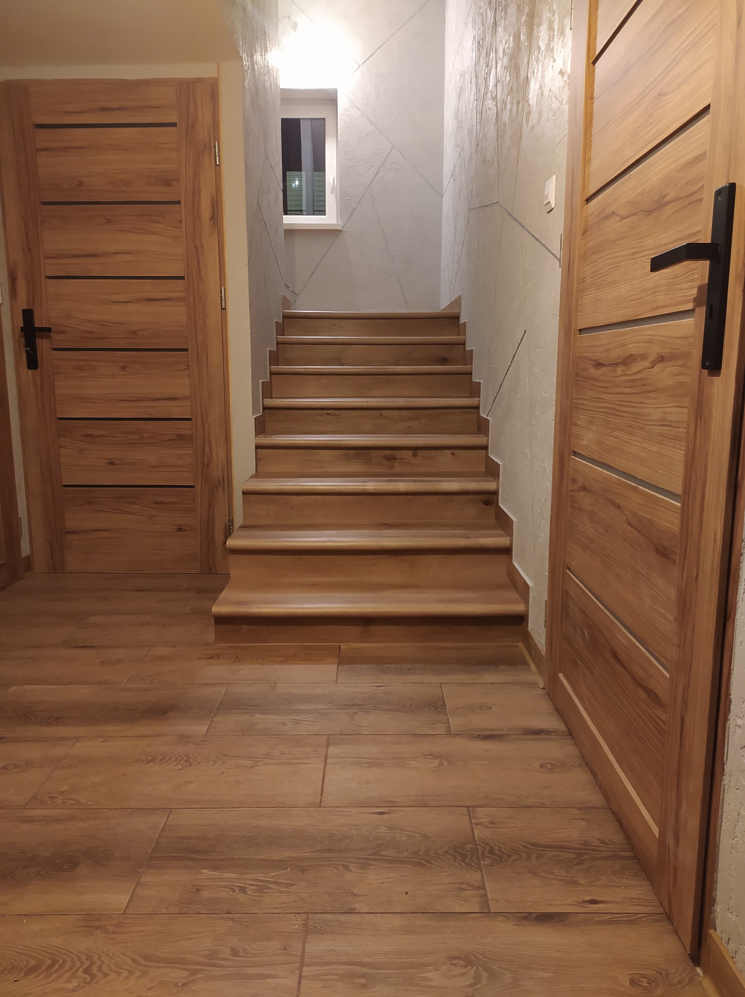 Klatka schodowa z drewnianymi schodami i panelami podłogowymi imitującymi drewno, dwie drewniane drzwi z czarnymi klamkami po bokach, ściana z dekoracyjną szarą fakturą i małe okno na podeście.