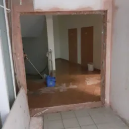 Otw&oacute;r po wyburzonej ścianie z widocznymi śladami prac murarskich, fragmentem instalacji i niebieskim wiadrem na brudnej podłodze.