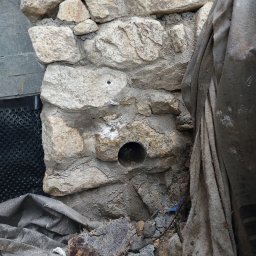 KRASH - Fragment kamiennej ściany z otworem na rurę, zabezpieczonej folią budowlaną i czarną membraną drenażową.