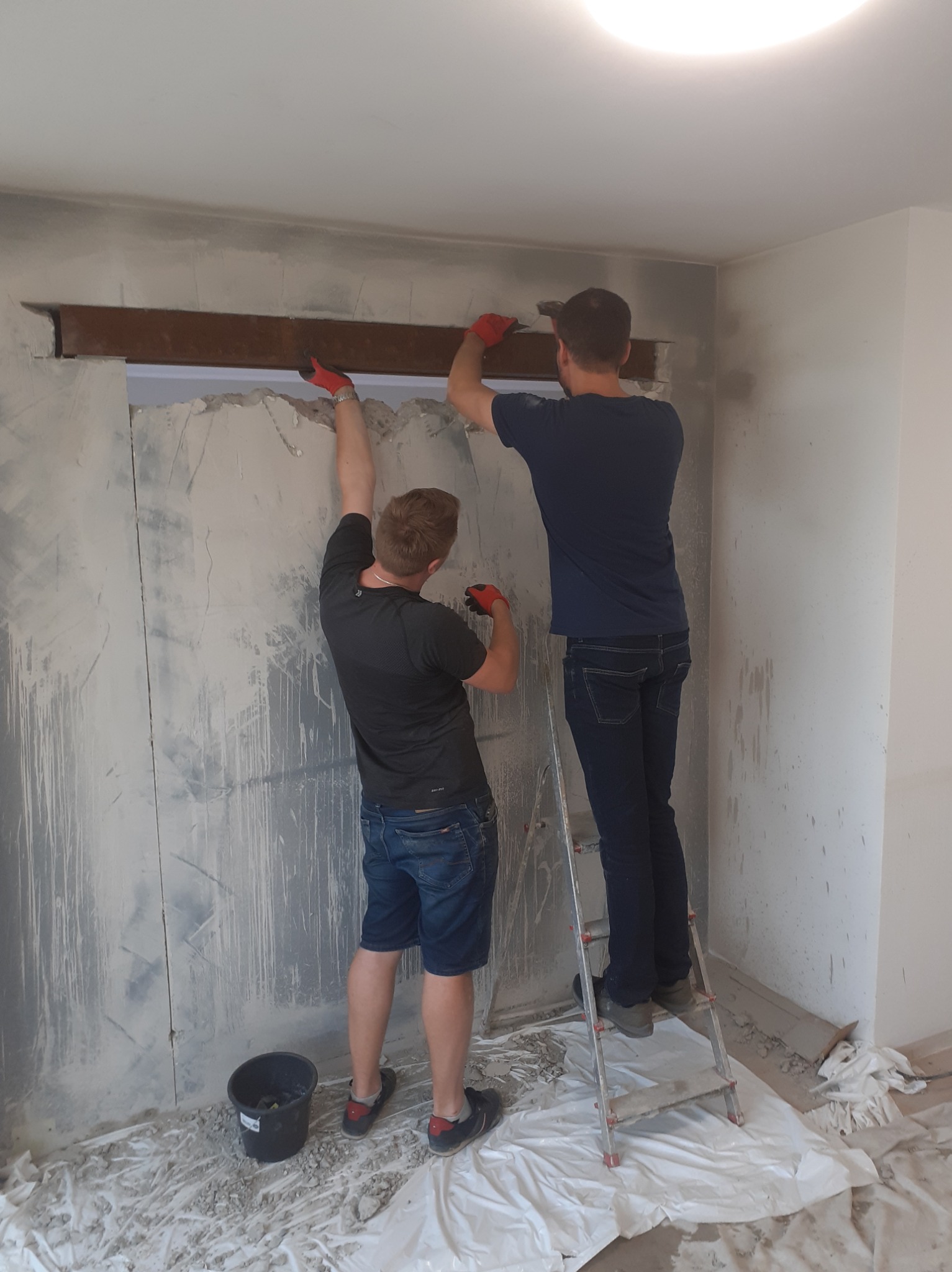 Dwóch mężczyzn w rękawicach ochronnych usuwa fragment ściany nadproża, wspierając się na drabinie, widoczne ślady po kuciu i gruz zabezpieczony folią.