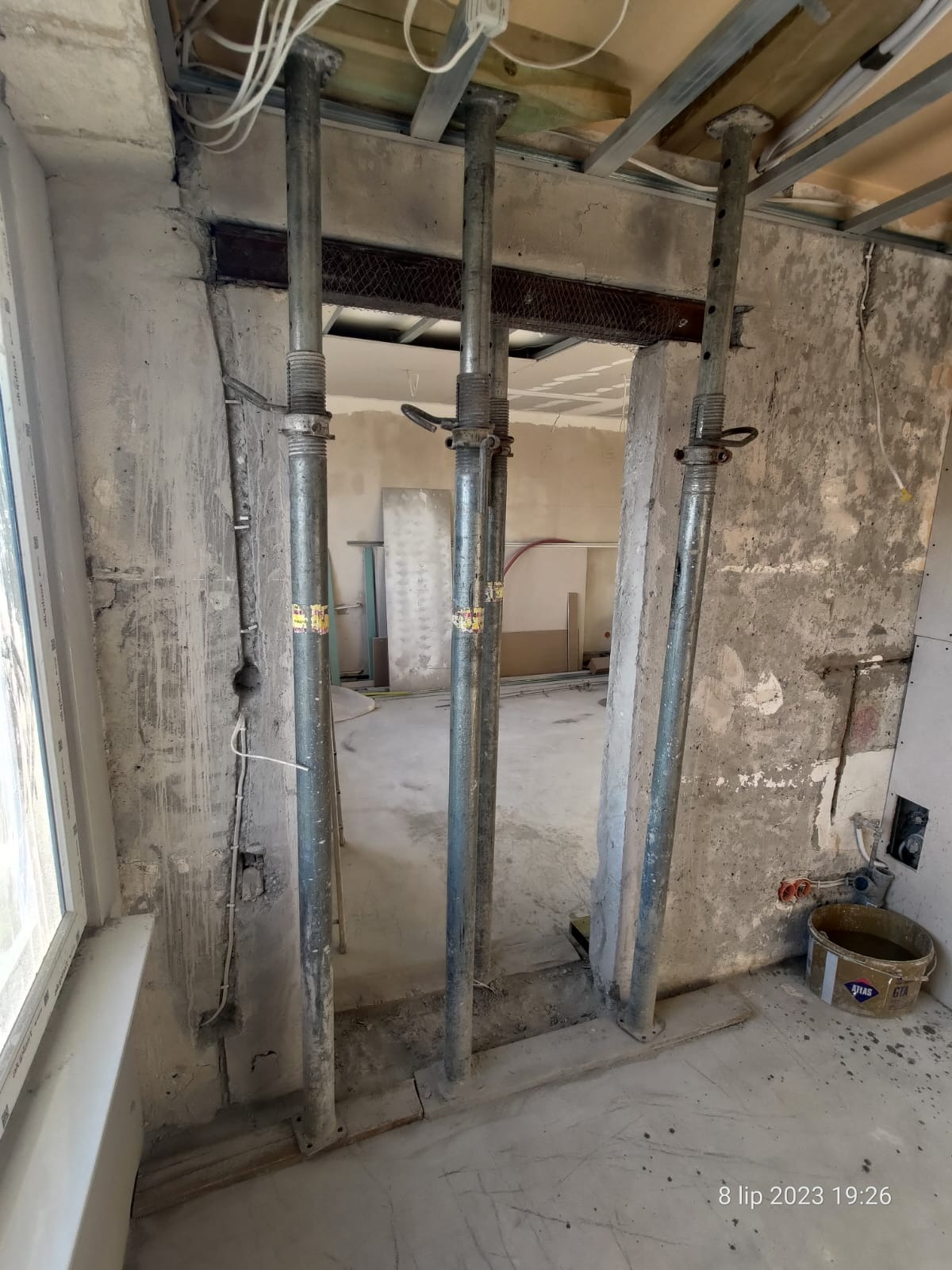 Wnętrze w trakcie prac budowlanych z trzema metalowymi stemplami podtrzymującymi strop, widoczne kable i resztki tynku na ścianach, wiadro z zaprawą w rogu.