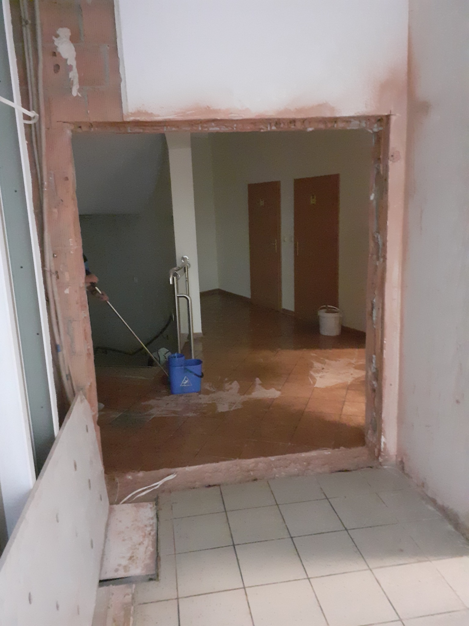 Otwór po wyburzonej ścianie z widocznymi śladami prac murarskich, fragmentem instalacji i niebieskim wiadrem na brudnej podłodze.