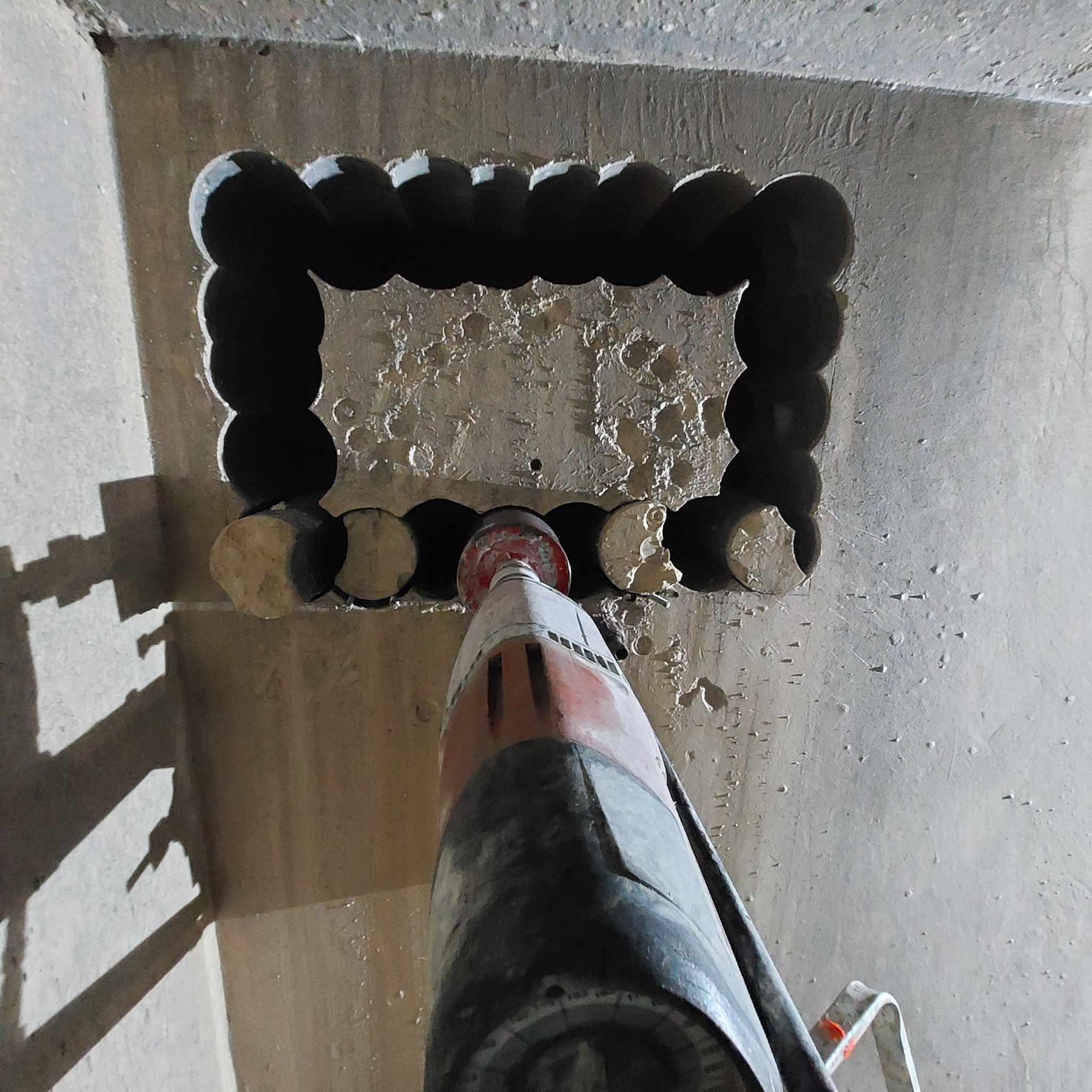 Wiercenie otworów w betonowej ścianie w celu wykonania otworu o kwadratowym kształcie. Widoczny wiertło rdzeniowe w trakcie pracy, a także pozostałości po wierceniu.