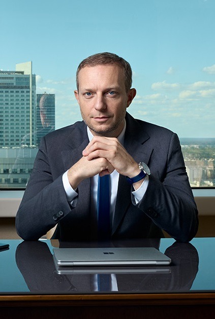 Portret mężczyzny w garniturze siedzącego za biurkiem z zamkniętym laptopem, widok na miasto w tle.