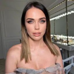 Portret kobiety z profesjonalnym makijażem, widoczne delikatne cienie na powiekach, podkreślone brwi i usta w odcieniu nude, w tle stanowisko do makijażu z lustrem podświetlanym żarówkami.