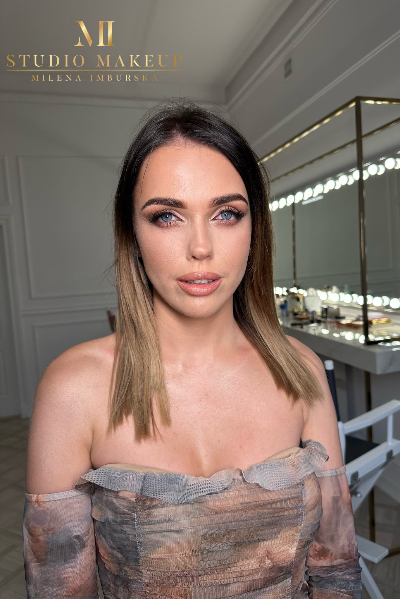 Portret kobiety z profesjonalnym makijażem, widoczne delikatne cienie na powiekach, podkreślone brwi i usta w odcieniu nude, w tle stanowisko do makijażu z lustrem podświetlanym żarówkami.