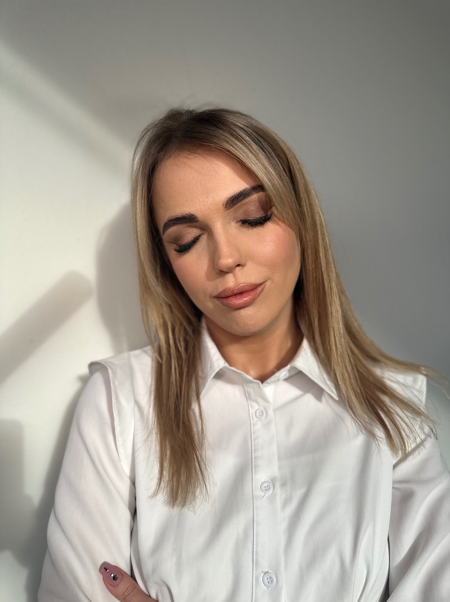 Portret kobiety z delikatnym, dziennym makijażem oczu i ust w odcieniach nude, ubrana w białą koszulę, z delikatnymi refleksami światła na twarzy i ścianie w tle.