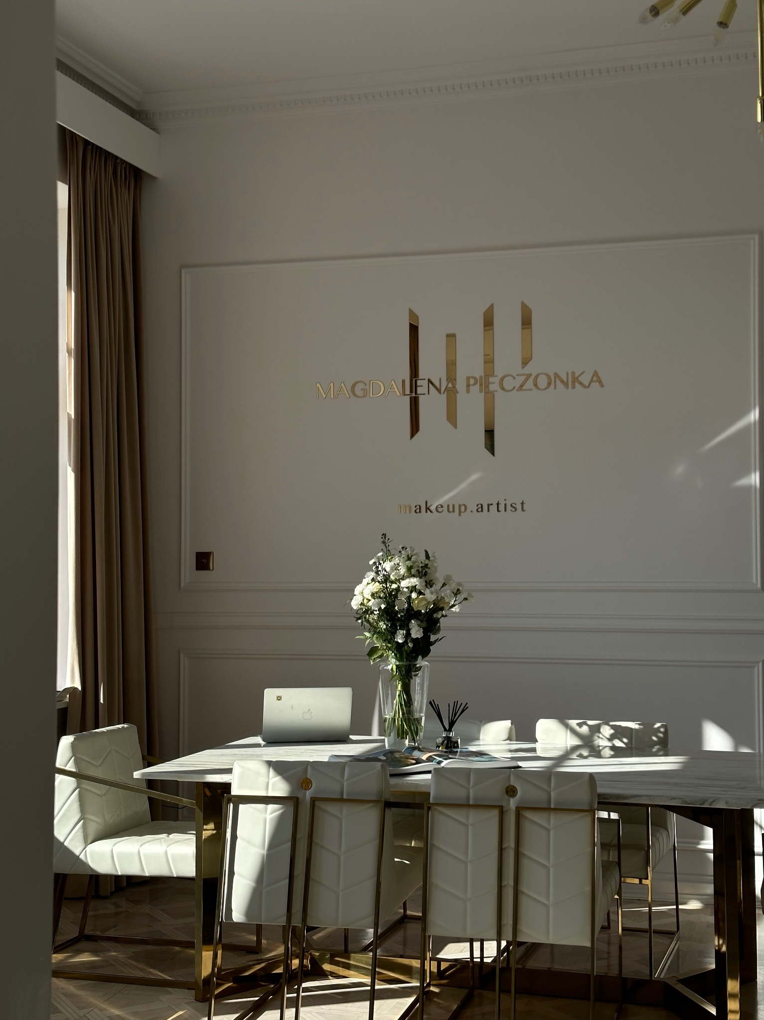 Eleganckie wnętrze studia z marmurowym stołem, białymi krzesłami z pikowaniem, laptopem, wazonem z kwiatami i złotym logo 'Magdalena Pieczonka makeup.artist' na ścianie w tle, oświetlone...