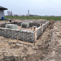 Adrian Betlej Usługi Remontowo Wykończeniowe - Wznoszenie fundamentów z bloczków betonowych na gruncie, widoczne palety z bloczkami, drewniane szalunki i odsłonięta ziemia w tle krajobraz polny.