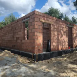 Murowany budynek w trakcie budowy z czerwonej cegły ceramicznej. Widoczne okna i drzwi. Fundament zabezpieczony folią kubełkową. W tle drzewa i błękitne niebo.