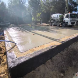 Wylany fundament pod dom jednorodzinny, zbrojenie widoczne, beton świeży, na tle lasu i betoniarki. Obrzeża zabezpieczone folią kubełkową.