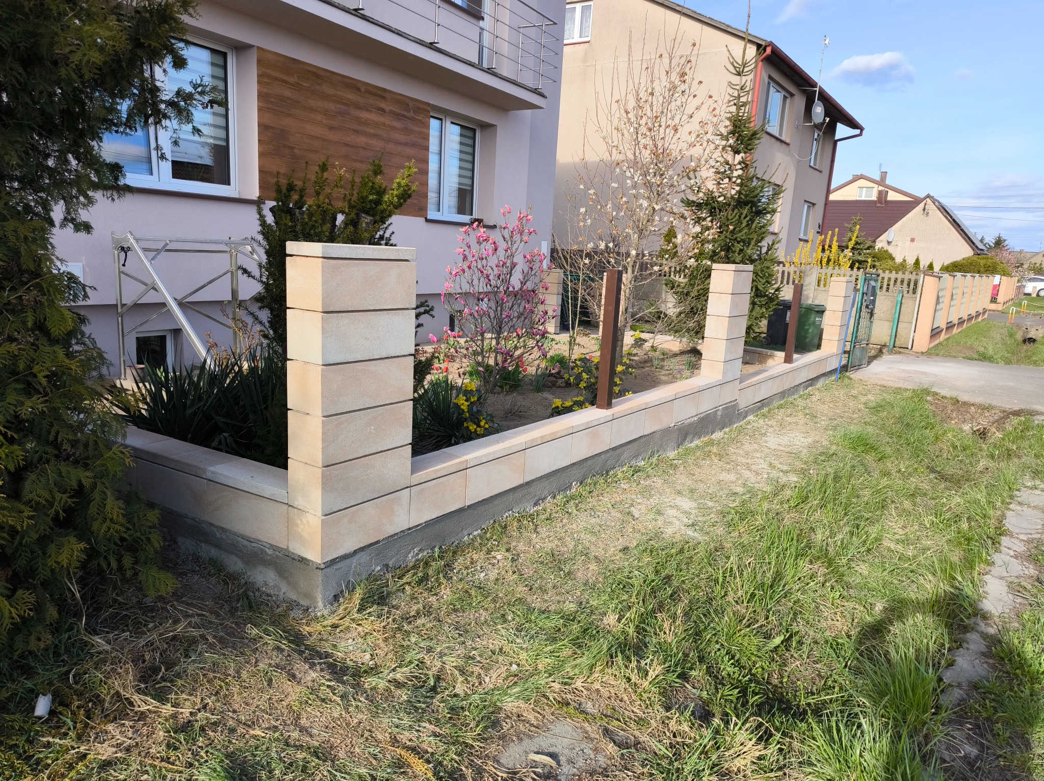 Nowoczesny, niski murek z beżowych, betonowych bloczków oddzielający ogródek od chodnika, z widocznymi nasadzeniami roślin i kwiatów, na tle dwukondygnacyjnego domu z elementami drewnianej elewacji.