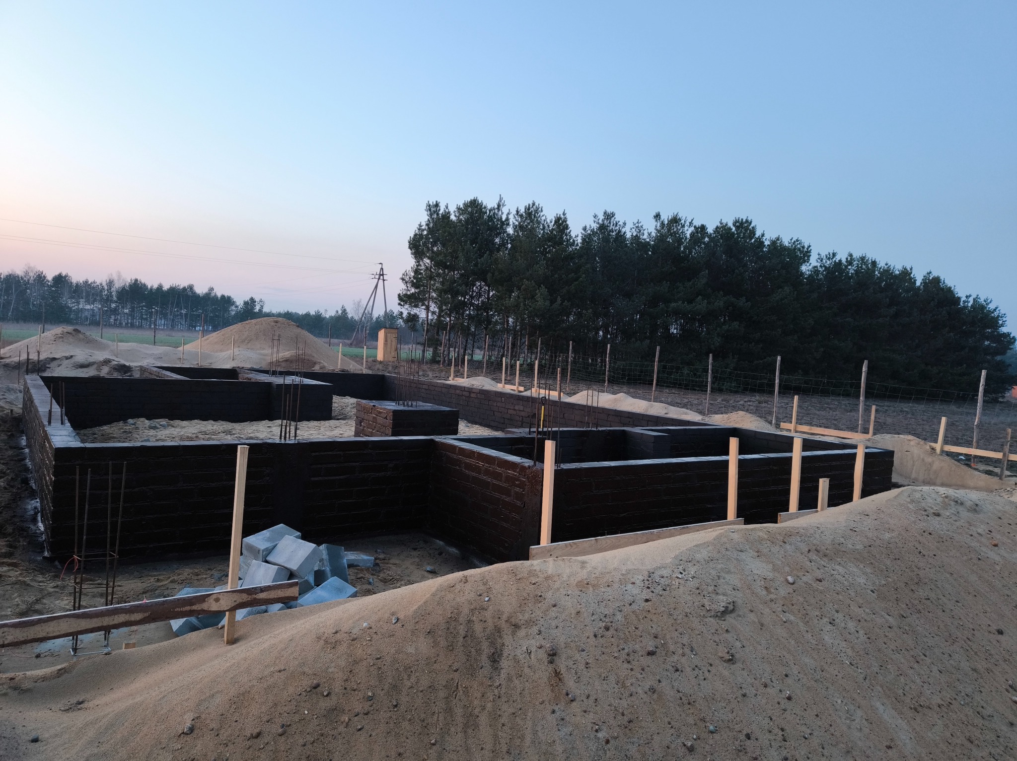 Wzniesione ściany fundamentowe z czarnej cegły, zabezpieczone przed wilgocią, otoczone piaskiem, z widocznymi fragmentami zbrojenia i drewnianych podpór, na tle drzew i nieba o zmierzchu.