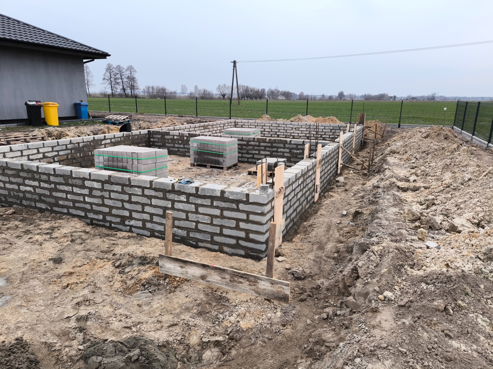 Wznoszenie fundamentów z bloczków betonowych na gruncie, widoczne palety z bloczkami, drewniane szalunki i odsłonięta ziemia w tle krajobraz polny.