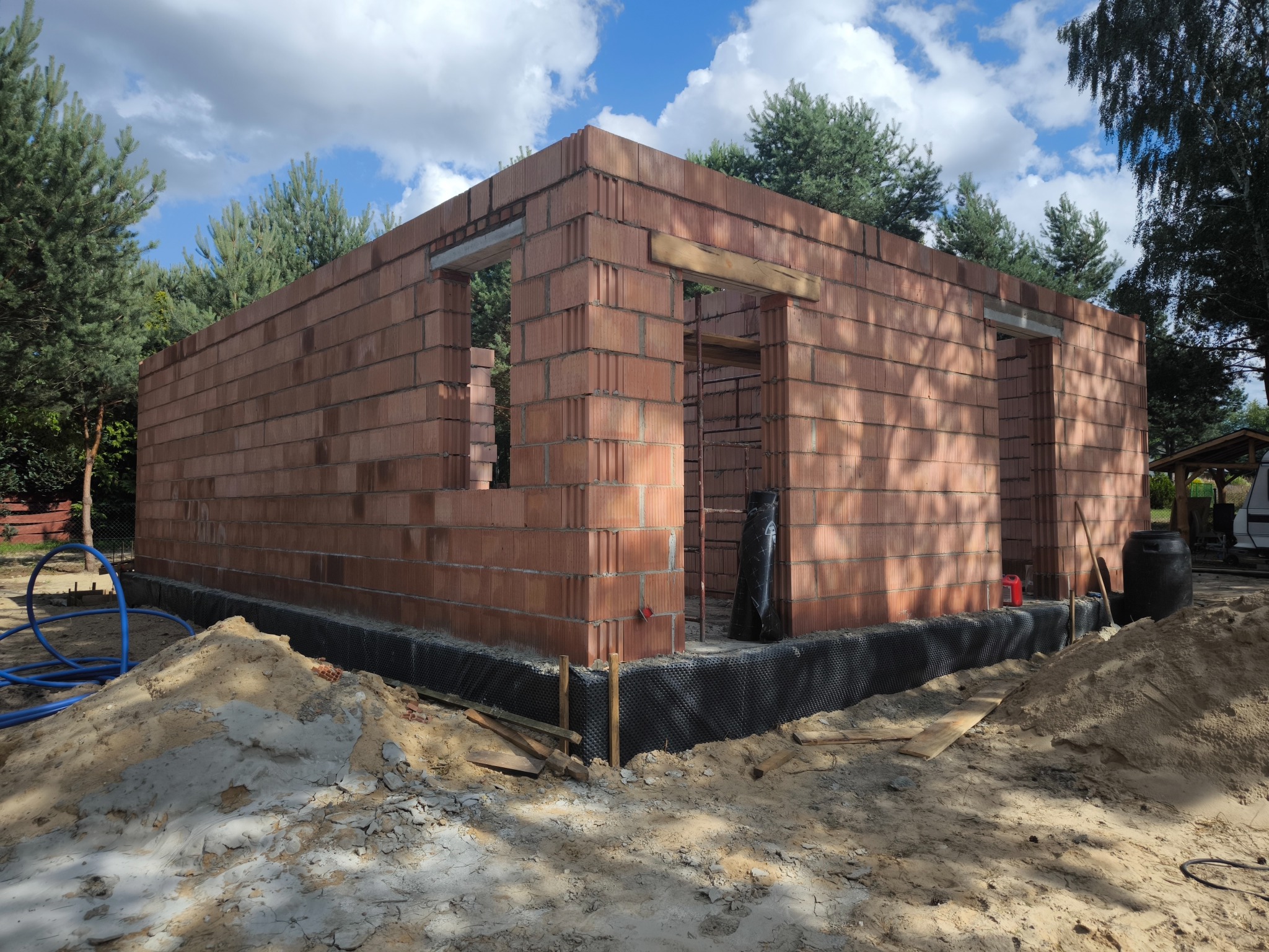 Murowany budynek w trakcie budowy z czerwonej cegły ceramicznej. Widoczne okna i drzwi. Fundament zabezpieczony folią kubełkową. W tle drzewa i błękitne niebo.
