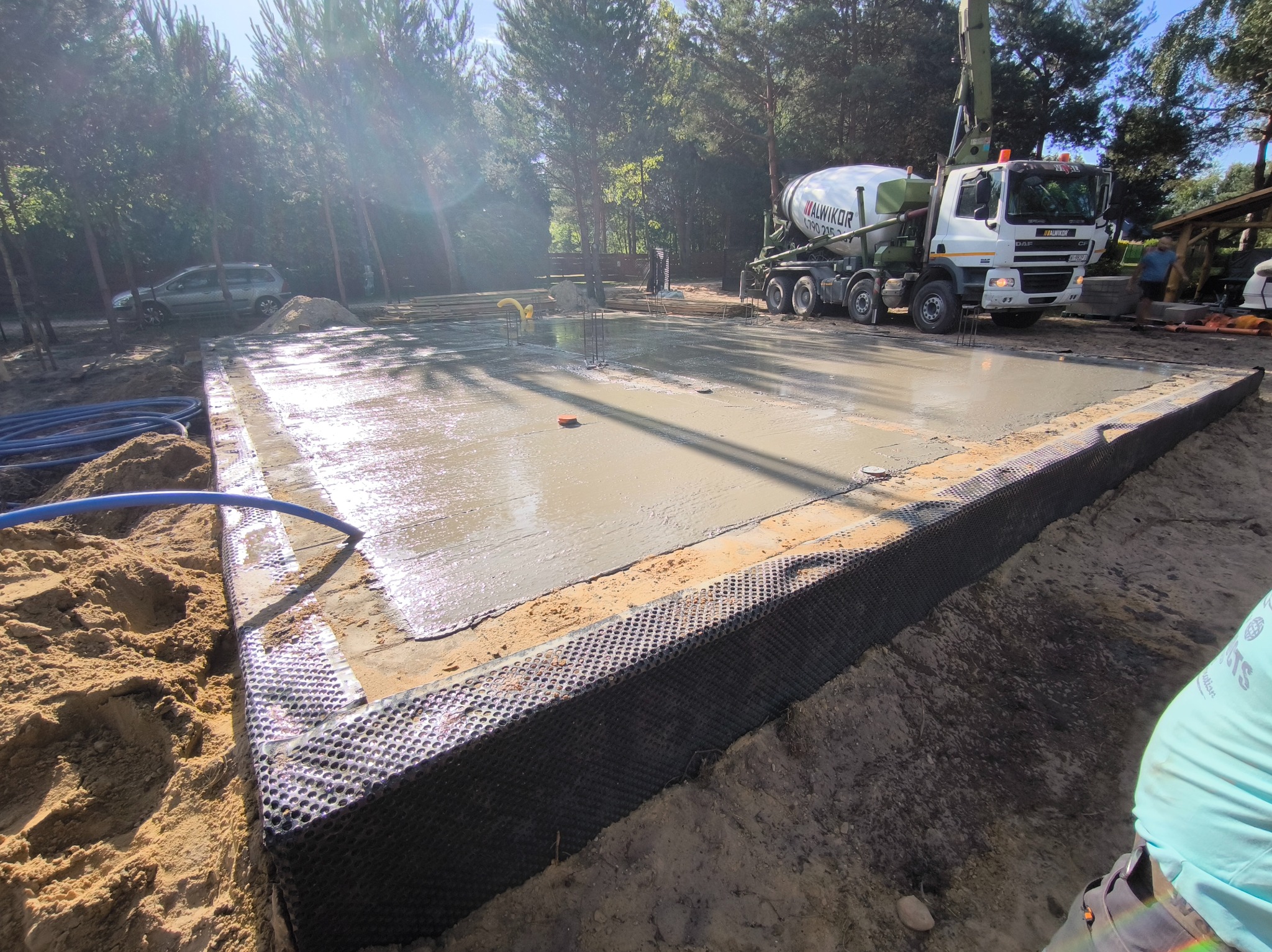 Wylany fundament pod dom jednorodzinny, zbrojenie widoczne, beton świeży, na tle lasu i betoniarki. Obrzeża zabezpieczone folią kubełkową.