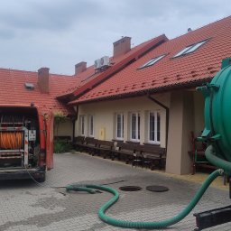 Kamwas Pogotowie kanalizacyjne - Pomarańczowy wąż zwinięty na bębnie w otwartym bagażniku czerwonego samochodu dostawczego, z zielonym wężem prowadzącym do kanalizacji i zielonym zbiornikiem na przyczepie w tle budynku z czerwoną...