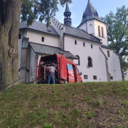 Kamwas Pogotowie kanalizacyjne - Pracownik w Gnojniku wyciąga wąż z czerwonego samochodu dostawczego zaparkowanego na wzgórzu przed białym kościołem z wieżami.