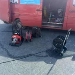 Sprzęt do czyszczenia rur kanalizacyjnych ustawiony przed otwartym busem z reklamą firmy oferującej usługi sanitarne, w tle zwinięte węże i narzędzia.