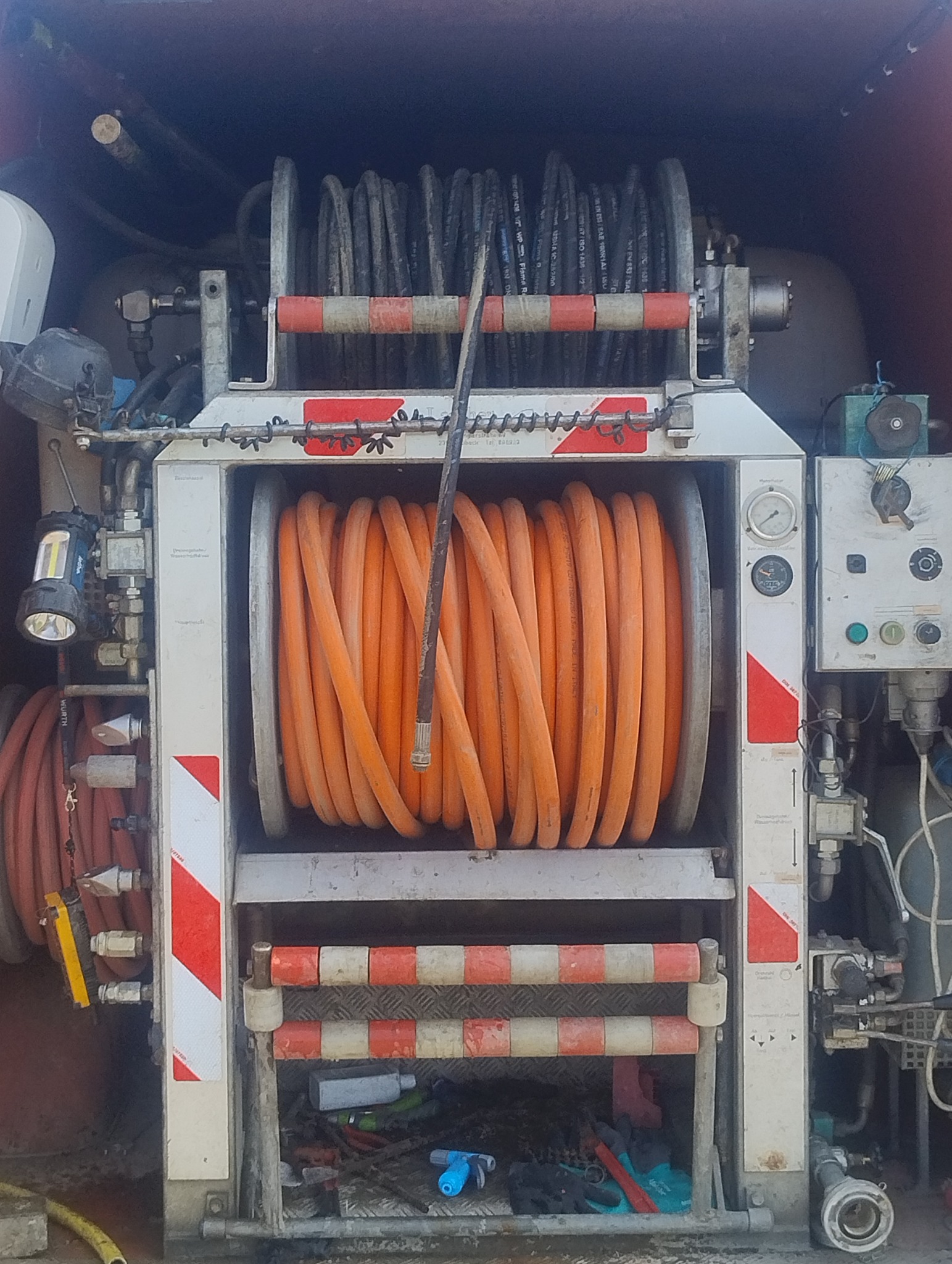 Pomarańczowy wąż zwinięty na bębnie wewnątrz pojazdu serwisowego z elementami ostrzegawczymi, widoczne narzędzia i aparatura hydrauliczna.
