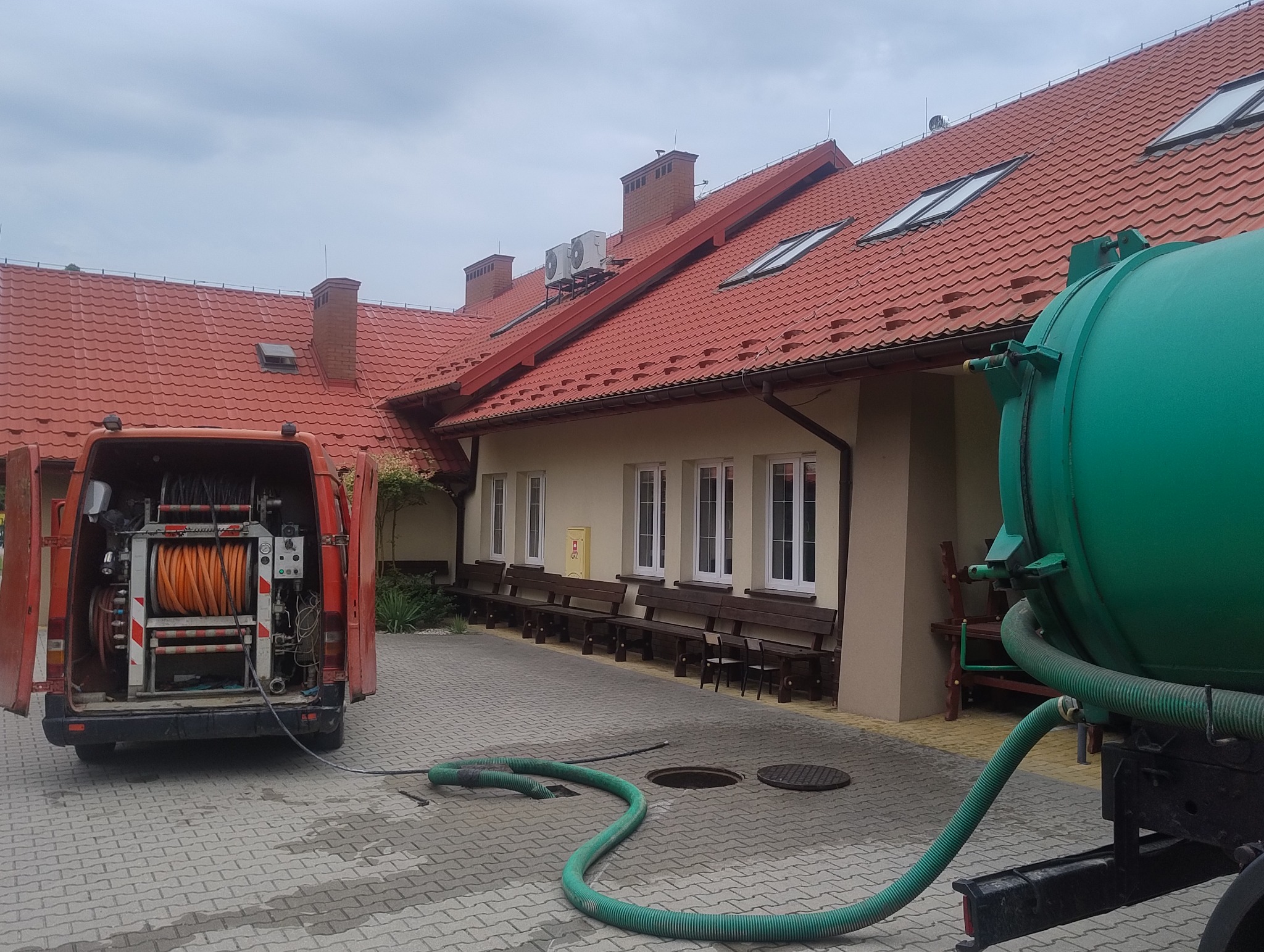 Pomarańczowy wąż zwinięty na bębnie w otwartym bagażniku czerwonego samochodu dostawczego, z zielonym wężem prowadzącym do kanalizacji i zielonym zbiornikiem na przyczepie w tle budynku z czerwoną...