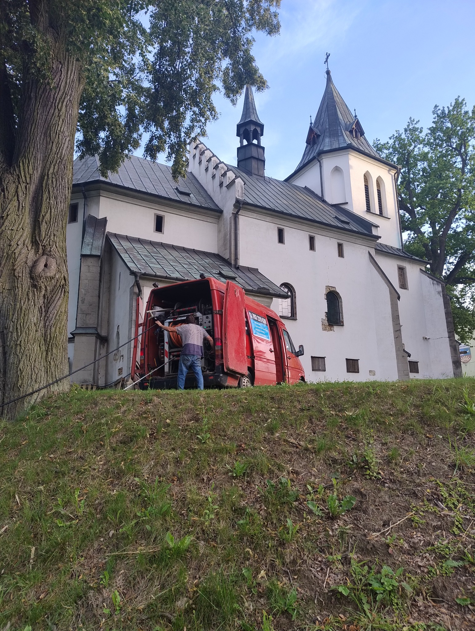 Pracownik w Gnojniku wyciąga wąż z czerwonego samochodu dostawczego zaparkowanego na wzgórzu przed białym kościołem z wieżami.