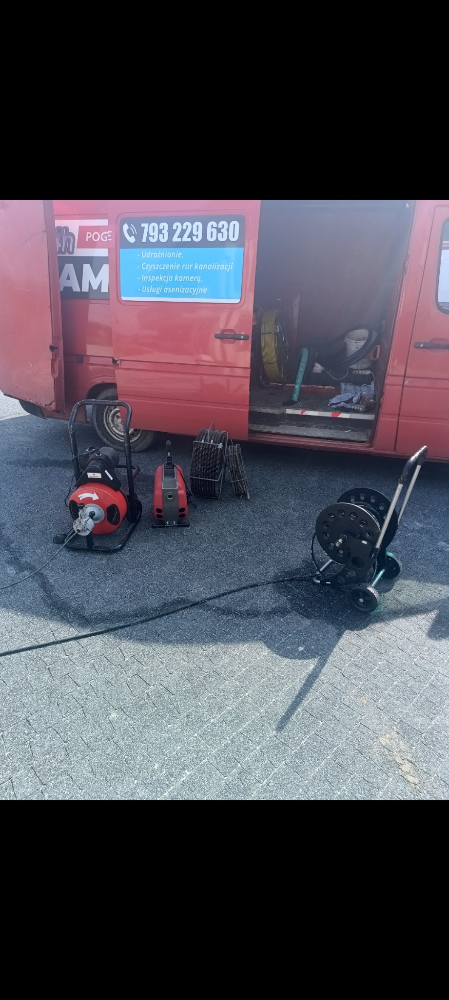 Sprzęt do czyszczenia rur kanalizacyjnych ustawiony przed otwartym busem z reklamą firmy oferującej usługi sanitarne, w tle zwinięte węże i narzędzia.